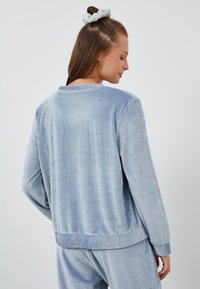 Jasnoniebieski sweter z kordurowej tkaniny z ściągaczami przy rękawach i dole. Charakteryzuje się luźnym krojem i subtelną fakturą. Widok z tyłu ukazuje detale dekoltu i przeszyć.
