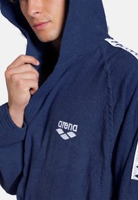Marineblå hætteklædte badekåbe lavet af frotté, forsynet med et broderet "arena" logo og mønstrede detaljer på ærmerne.