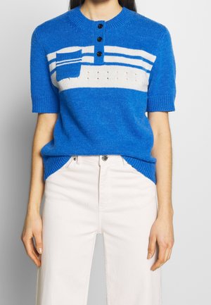 Persoon draagt een blauw gebreid topje met korte mouwen, witte strepen, een klein zakje en zwarte knopen, gecombineerd met witte broeken met hoge taille.
