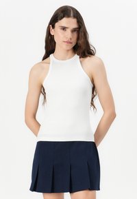 Weißes, geripptes Tanktop mit einem Halterneck-Design, das eine enge Passform und eine glatte Textur bietet. Kombiniert mit einem dunkelblauen, plissierten Rock.