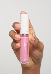 Plumper per labbra in un tubo trasparente con liquido rosa, riflessi argentati e tappo bianco; tenuto in una mano con unghie naturali e non smaltate.
