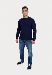 Camicia a maniche lunghe blu navy in tessuto leggero, design aderente, abbinata a jeans in denim blu e sneakers scure. Piccolo dettaglio del logo sulla camicia.