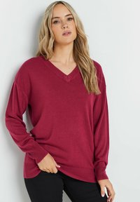 Bordeauxrotes V-Ausschnitt Sweatshirt mit langen Ärmeln, gestrickter Textur, geripptem Saum und Bündchen sowie lockerer Passform, kombiniert mit schwarzen Hosen.