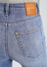 Jeans de mezclilla azul claro con un parche de cuero en la cintura, con costuras clásicas, un bolsillo trasero y un corte estructurado.