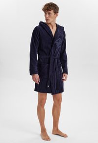 Navyblaue Baumwollbademantel mit Kapuze, vorne mit einer Bindung, zwei Taschen und mit einer strukturierten Frottee-Oberfläche. Logo auf der Brust gedruckt.