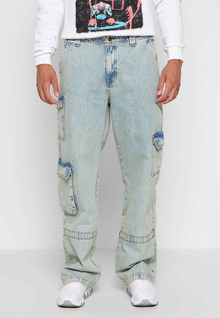 Pantaloni cargo in denim azzurro chiaro con vestibilità ampia, molteplici tasche laterali e un effetto sbiadito. Indossati con una maglietta bianca a maniche lunghe con grafiche.
