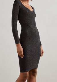 Abito nero a maglia a coste con maniche lunghe e scollatura a V; presenta una vestibilità aderente e una leggera lucentezza su tutta la stoffa.