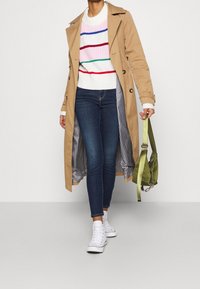 Abrigo trinchera beige con forro a cuadros, suéter blanco con rayas de colores, jeans ajustados oscuros y zapatillas blancas. Llevando un bolso verde.
