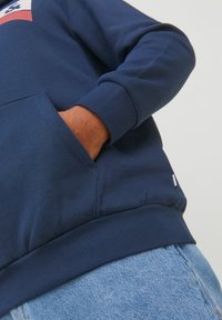 Sudadera azul marino con capucha, bolsillo frontal y puños acanalados. Hecha de una tela suave con un detalle de logo en el pecho y una pequeña etiqueta en el dobladillo.