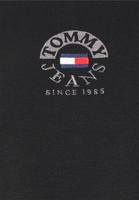 Schwarzer Stoff mit gesticktem Tommy Jeans-Logo, das eine rot-weiße-blau Flagge und den Text "Seit 1985" zeigt.