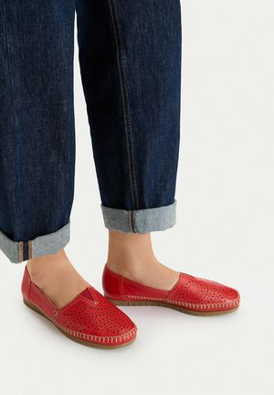 Zapatos de piel rojos sin cordones con diseño perforado, usados con vaqueros azul oscuro remangados y pies descalzos sobre un fondo liso.