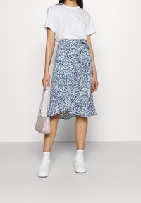 Blommig blå midikjol med volangnederkant, tillsammans med en vit t-shirt och vita sneakers. Håller en liten rosa handväska.