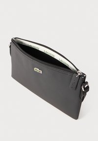 Pochette nera con texture e chiusura a zip, caratterizzata da un accento del logo e un rivestimento interno a contrasto con marchi ripetuti. Include una tracolla regolabile.