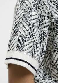 Adītais īsās piedurknes svīteris baltā un tumši zilā krāsā ar zigzakveida rakstu. Iekļautas ribotas aproces ar tumši zilu svītru. Tekstūrveida audums.