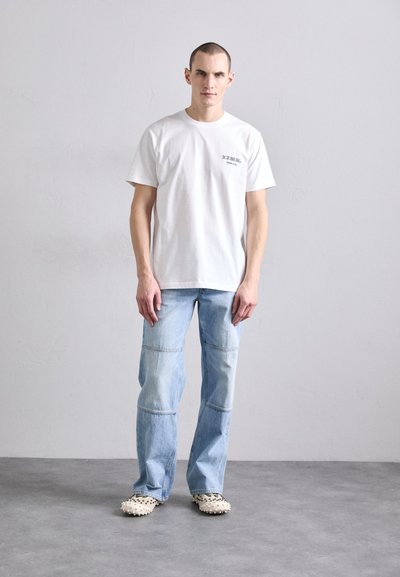 T-shirt en coton blanc avec un petit logo noir, associé à un jean en denim bleu clair avec un design en patchwork, et des sabots à motifs.