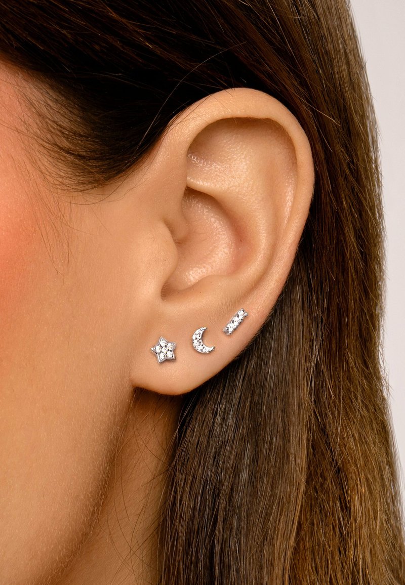 Pendientes de plata con tachuelas que presentan una estrella, una luna creciente y una barra recta, decorados con piedras preciosas transparentes, sobre un fondo neutro.