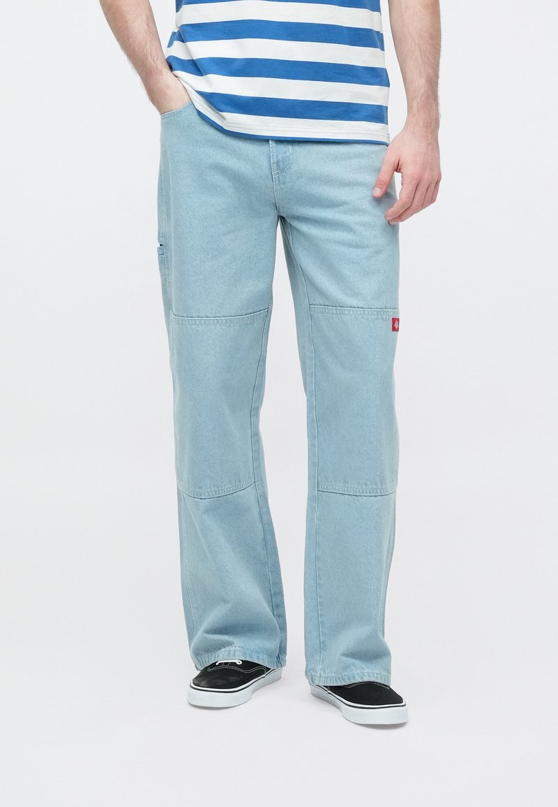 Hellblaue Cargo-Hose mit lockerem Schnitt, seitlichen Taschen und einem kleinen roten Logostreifen. Kombiniert mit schwarzen Schuhen und einem gestreiften Oberteil.