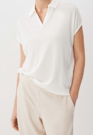 Femme portant une chemise blanche texturée à manches courtes avec col et un pantalon beige clair, une main dans la poche, sur un fond neutre.