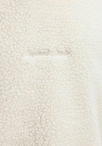 Crème-kleurige fleece stof met een gestructureerd oppervlak met kleine, verhoogde bobbels. Bevat de reliëftekst "Original's Studio."