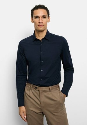 Navy long-sleeve button-up shirt with a tailored fit, smooth texture, and light sheen. Paired with beige trousers, featuring side pockets.
