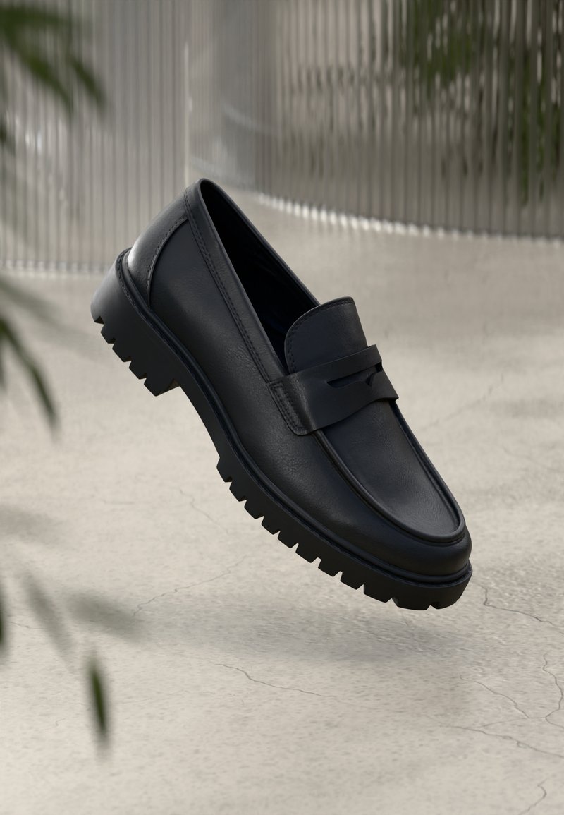 Mocassin en cuir noir avec semelle crantée épaisse, détail bride penny, sur un fond neutre texturé avec des plantes floues.