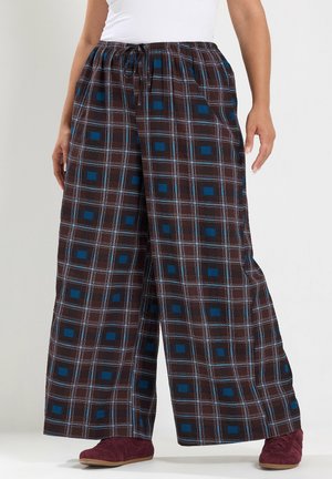 Personne portant un pantalon large marron à carreaux avec des carrés bleus et des chaussures bordeaux, debout sur un fond clair uni.