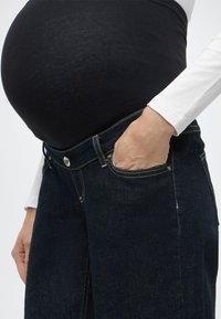 Τζιν εγκυμοσύνης με βαθύ μπλε τελείωμα τζιν και ραμμένες λεπτομέρειες, με ελαστική μαύρη επάνω στρώση και διακοσμητικές λεπτομέρειες στις μπροστινές τσέπες.