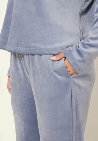 Mjuk ljusblå sammet loungedress med långa ärmar, byxor med elastisk midja, och en hand avslappnat placerad i fickan.