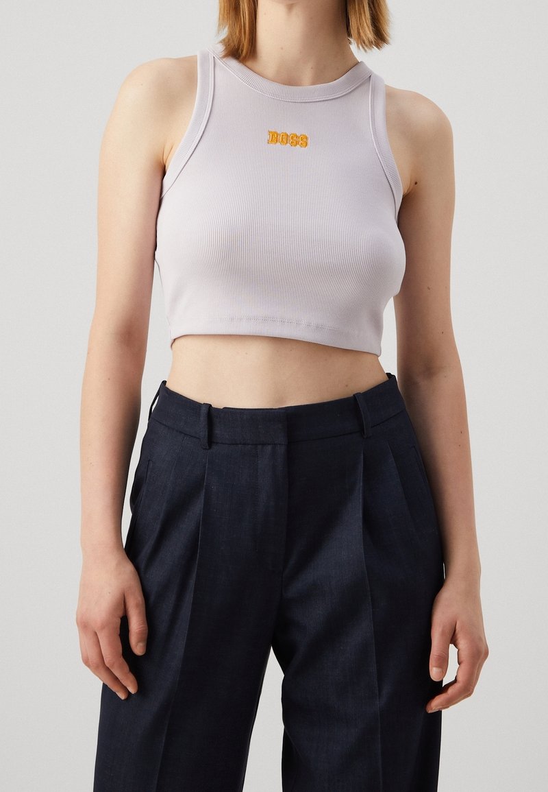 Femme portant un débardeur court côtelé blanc avec "BOSS" en lettres orange et un pantalon ample taille haute foncé.