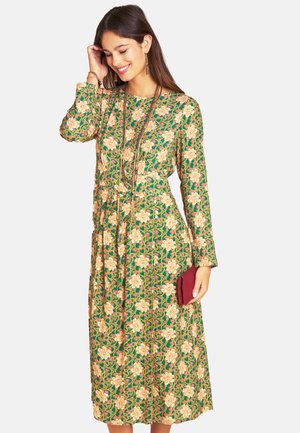 Woman wearing a green, long-sleeve floral dress holding a red clutch, smiling and touching her hair, with long necklaces and earrings.