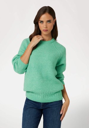 Maglione in maglia verde menta con texture a costine, scollo rotondo e maniche a palloncino; abbinato a jeans blu, la modella posa con una mano appoggiata sulla spalla.