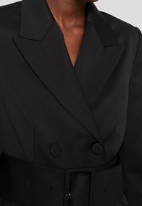 Blazer negro de doble botonadura con solapa ancha, que presenta una cintura ceñida con cinturón, tejido texturizado y botones redondos para el cierre.