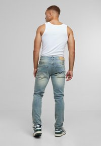 Hombre de pie de espaldas, vistiendo una camiseta sin mangas blanca, jeans ligeros azules y zapatillas Nike en blanco y negro, sobre un fondo liso.