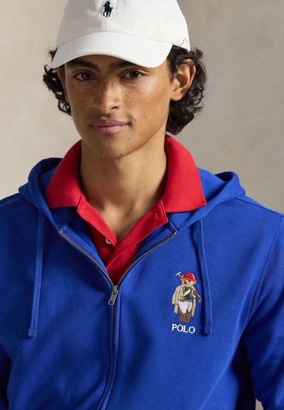 Modrá mikina na zip s červenou košilí s límečkem pod ní. Na mikině je vyšitý medvědí znak a nápis "POLO". Oblečení doplňuje bílá čepice.