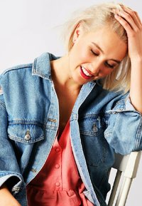 Lysblå denimjakke med slidte detaljer og sølvhardware, lagdelt over en pink kjole med knapper og en slået talje.