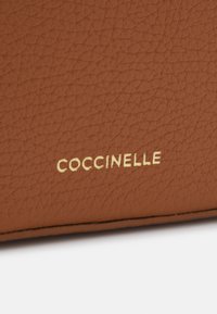 Smeđi kožni novčanik s teksturiranom površinom, s zlatnim reljefnim logotipom "COCCINELLE" na donjem rubu. Minimalistički dizajn.