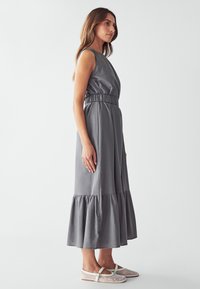 Vestido gris sin mangas con cintura elástica y dobladillo con volantes, hecho de una tela suave. Combinado con zapatos planos de malla blanca. Fondo neutro.