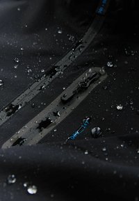 Vodootporno crno jakna tkanina s vidljivim kapima vode, s džepom na patentni zatvarač i teksturiranim mrežastim uzorkom. Plavi naglasak na patentnom zatvaraču.