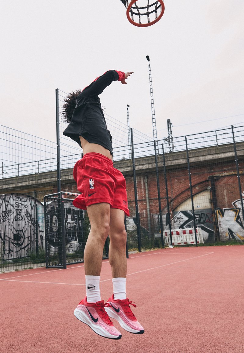 Koszykarz w powietrzu, ubrany w czarną koszulkę, czerwone spodenki i czerwone buty Nike z białymi akcentami, na różowym boisku z tłem w graffiti.