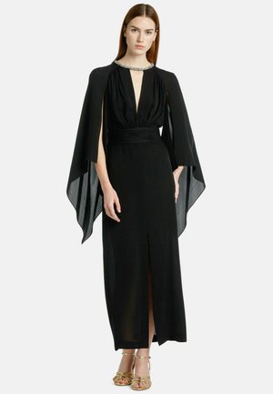 Zwarte avondjurk met diepe V-hals, vloeiende capesleuven en een zijsplit. Versierde halslijn en aansluitende taille, gecombineerd met gouden sandalen.