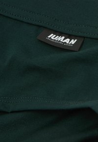 Ciemnozielony materiał bawełnianej koszulki, z naszytą metką z napisem "JUMAN" oraz "intimissimi uomo" w białym kolorze. Gładka struktura, bez widocznych wzorów.