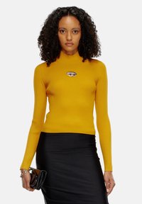 Maglione a collo alto a coste color senape con maniche lunghe, caratterizzato da un accento metallico del logo. Abbinato a una gonna nera aderente e una borsa.