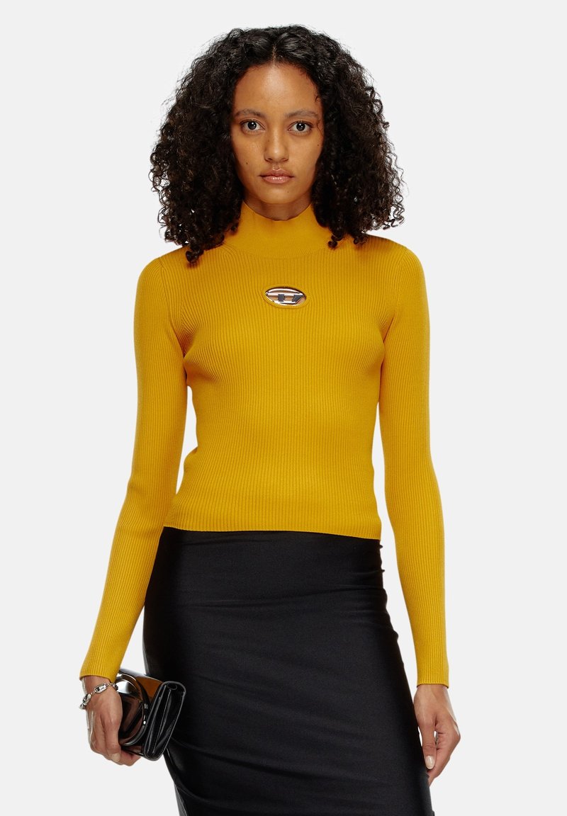 Maglione a collo alto a coste color senape con maniche lunghe, caratterizzato da un accento metallico del logo. Abbinato a una gonna nera aderente e una borsa.