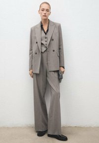 Un traje gris a medida que cuenta con un blazer de doble botonadura, pantalones de pierna ancha y un chaleco ajustado, combinado con una camisa negra y accesorios.