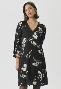 Robe noire avec un décolleté en V, des manches bouffantes 3/4 et un motif floral blanc. Conçue en tissu léger, elle offre une coupe ample et une longueur au genou.