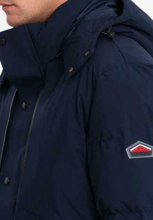 Giacca imbottita blu navy con colletto alto, bottoni a pressione e cappuccio con cordino. Presenta una patch con logo sulla manica sinistra. Tessuto liscio e texturizzato.