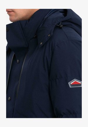 Giacca imbottita blu navy con colletto alto, bottoni a pressione e cappuccio con cordino. Presenta una patch con logo sulla manica sinistra. Tessuto liscio e texturizzato.