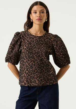 Frau mit brauner Leopardenmuster-Bluse mit Puffärmeln, dunklen Jeans, goldenen Creolen und einer klobigen goldenen Halskette vor einem schlichten Hintergrund.