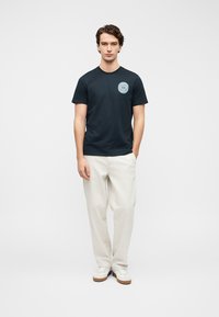 Μπλε βαμβακερό μπλουζάκι ναυτικού τύπου με κυκλικό λογότυπο στο αριστερό μέρος του στήθους, σε συνδυασμό με ανοιχτό μπεζ παντελόνι και λευκά αθλητικά παπούτσια.