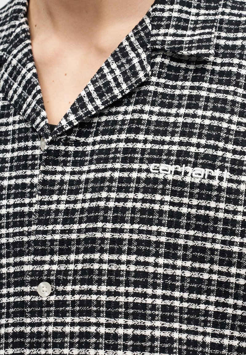 Primo piano del colletto di una camicia a bottoni a quadri bianco e nero con il logo "Carhartt" ricamato sul petto.